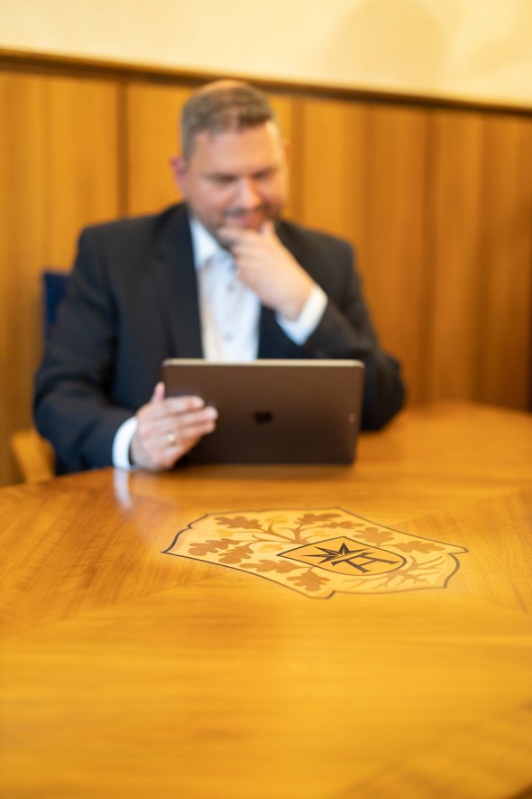 Bürgermeister Lambion sitzt an einem Holztisch auf dem das Wappen der Stadt Bad Arolsen ist und blickt auf ein Tablet