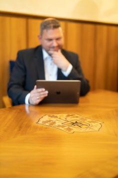 Wappen Stadt Bad Arolsen Bürgermeister Lambion sitzt an einem Holztisch auf dem das Wappen der Stadt Bad Arolsen ist und blickt auf ein Tablet