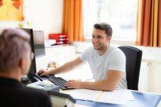 Bürgergespräch Eine freundliche Mitarbeiterin der Stadtverwaltung Bad Arolsen sitzt lächelnd hinter einer Plexiglasscheibe am Empfangstresen in der zentralen Anmeldung. Die Mitarbeiterin spricht mit einer Besucherin, die ihr gegenübersitzt.