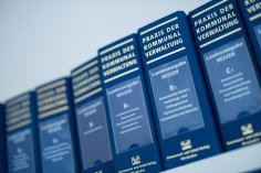 Eine Reihe von blauen Ordnern mit der Aufschrift 'Praxis der Kommunalverwaltung', Ausgabe Hessen, im Regal. Die Ordner behandeln verschiedene Themen der kommunalen Verwaltung, wie die Landkreisordnung, kommunales Kassen- und Rechnungswesen sowie Datenschutz.