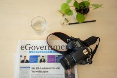 Ein Schreibtisch mit einer Zeitung mit der Überschrift 'eGovernment – Verwaltung digital', einer schwarzen Kamera mit Trageriemen, einem Glas Wasser, einem Bleistift, einem Anspitzer und einer kleinen Pflanze.